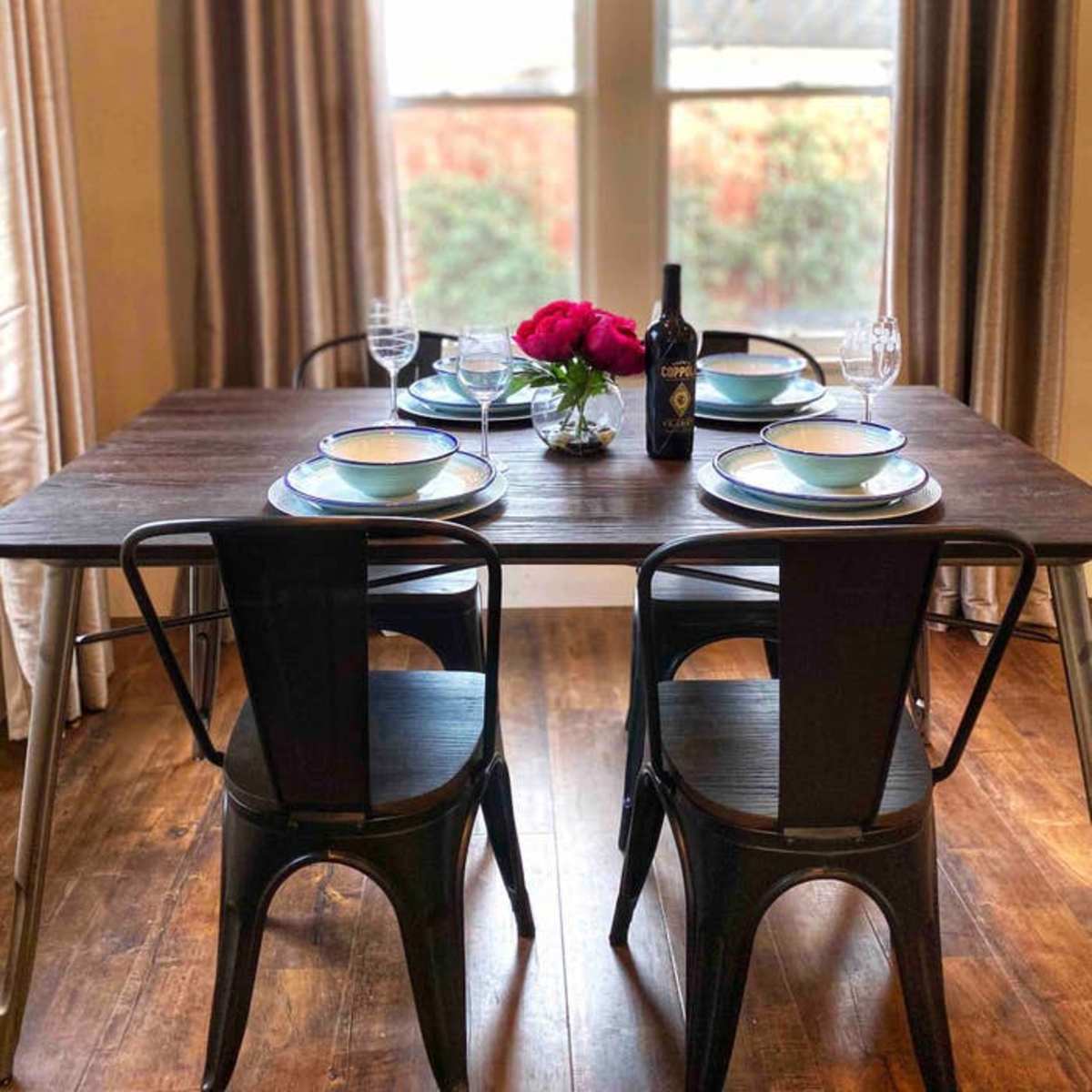 A dining table is set with plates and glasses, featuring a vase of flowers and a bottle of wine, positioned near a window with curtains.