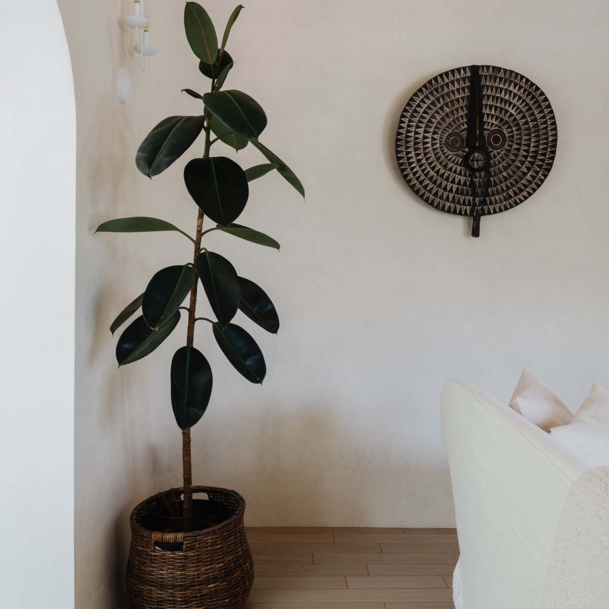 A potted plant stands in a woven basket beside a wall featuring a decorative mask mounted on it.