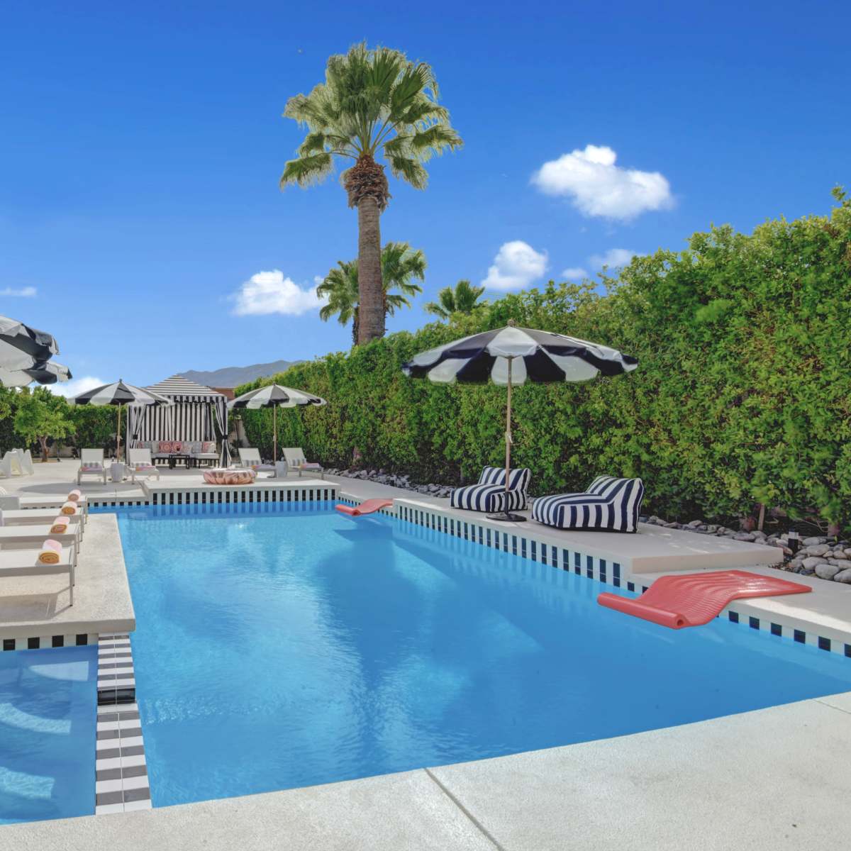 A rectangular swimming pool is surrounded by lounge chairs and striped umbrellas, with palm trees and a hedge in the background.