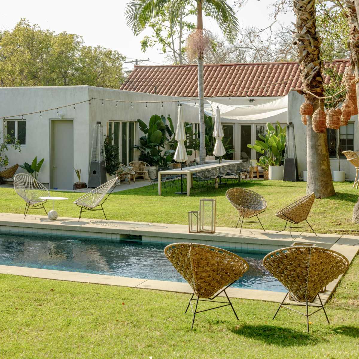 A modern house with a red-tiled roof is set next to a pool surrounded by lounge chairs and tropical greenery.