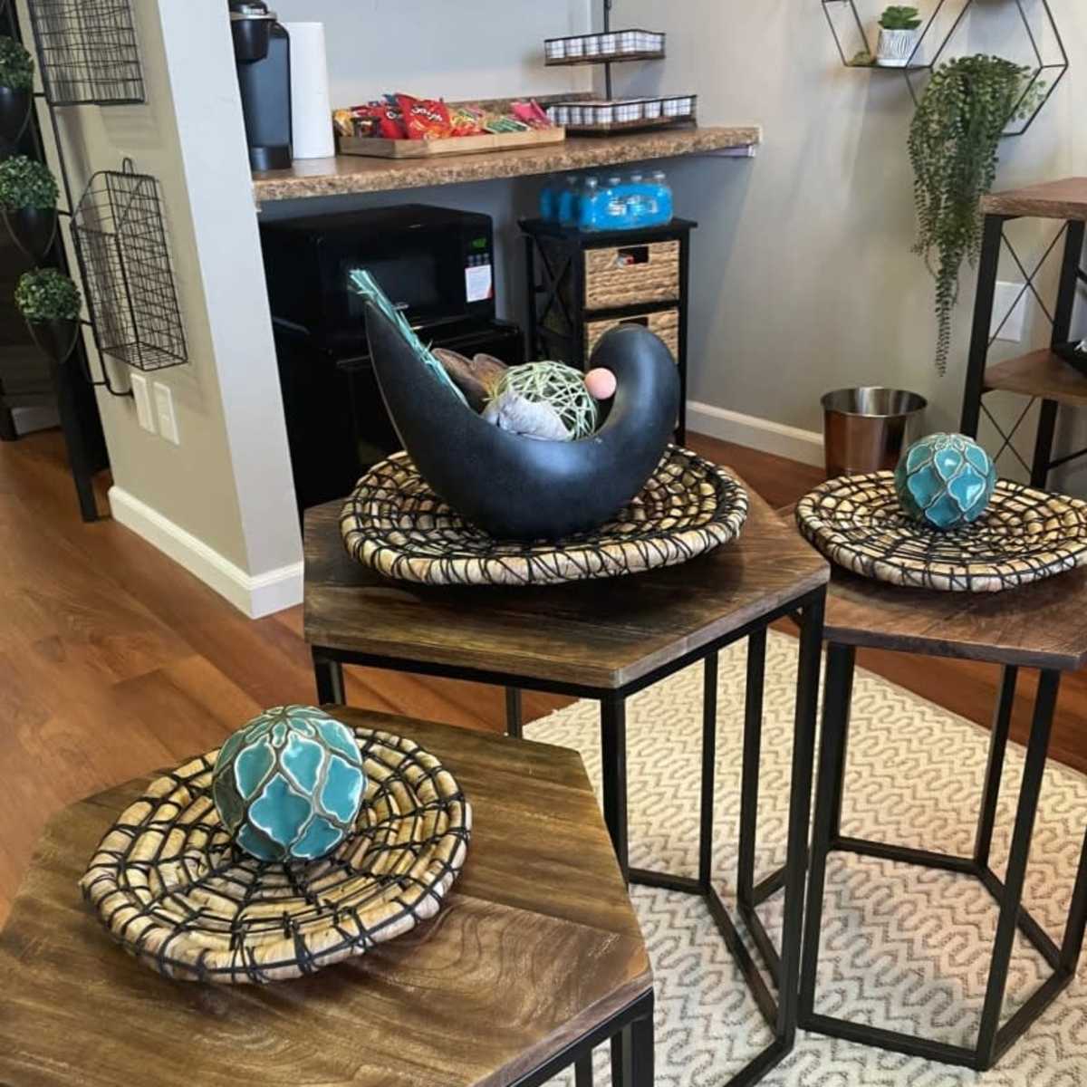 Three nested wooden tables of varying heights, each topped with decorative woven mats and ornamental objects, set in a modern room with plants and shelves in the background.