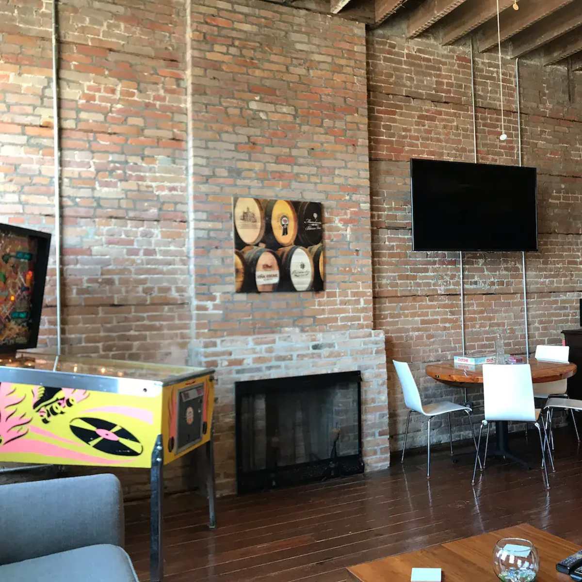 An interior space with exposed brick walls, a wooden floor, a round dining table with white chairs, a wall-mounted television, and a vintage arcade machine.