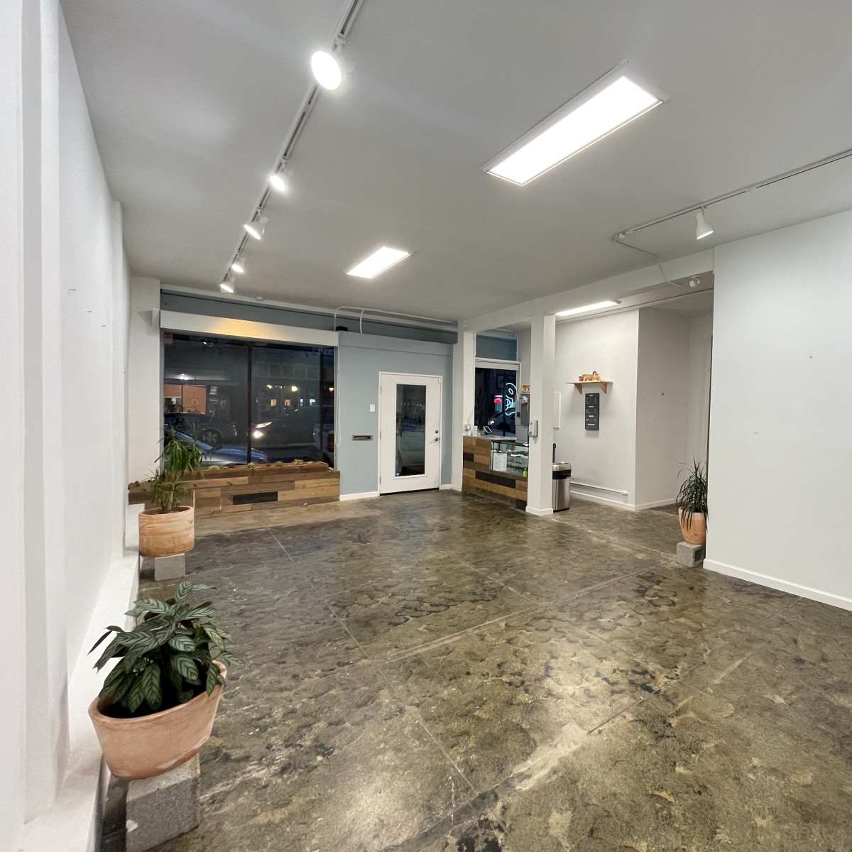 An empty interior space with polished concrete flooring, white walls, and a large window, featuring a door and a service counter in one corner.