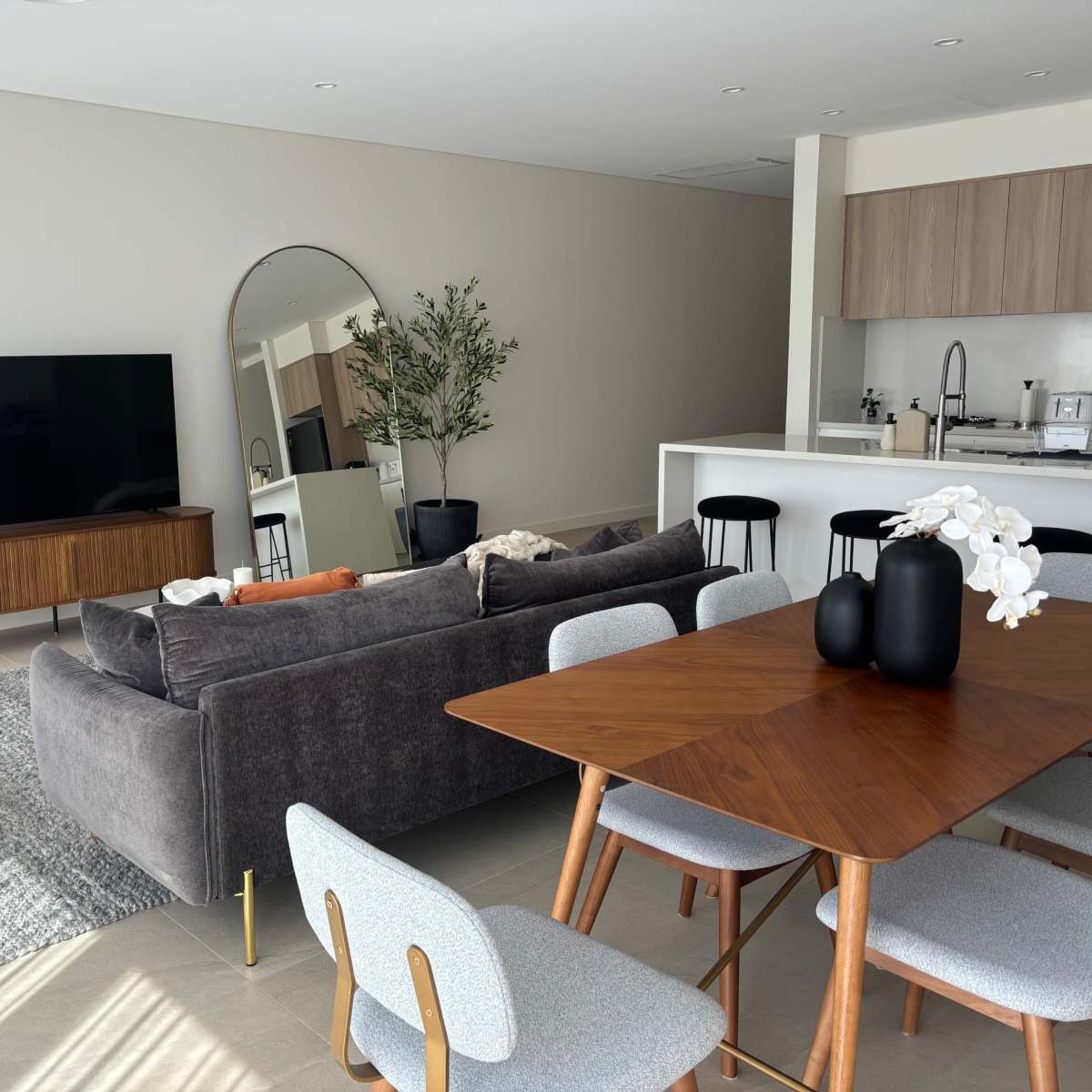 A modern living space featuring a gray sofa, a wooden dining table with chairs, a large television, and a kitchen area in the background.