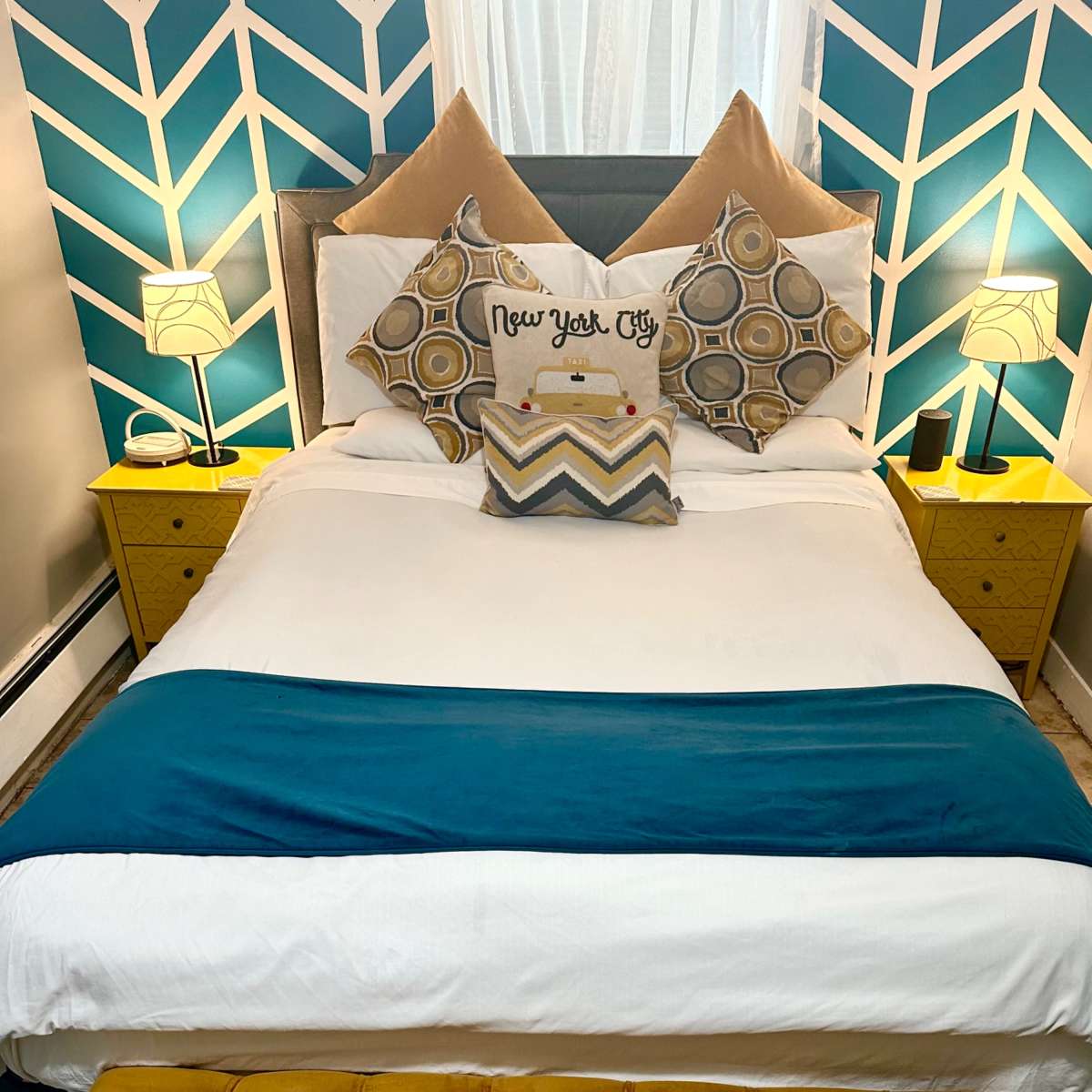 A neatly made bed with a blue accent strip, decorated with patterned pillows, flanked by two yellow nightstands and a mustard-colored bench.