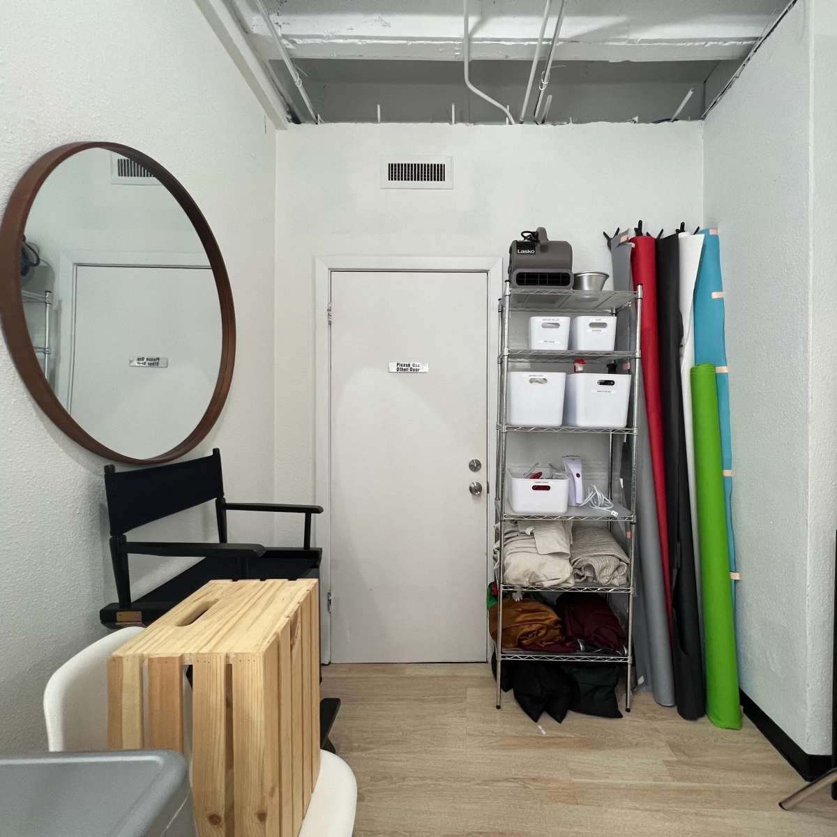 A small studio room with a round mirror, a door, a shelving unit filled with storage boxes, and rolls of colored paper stacked against the wall.
