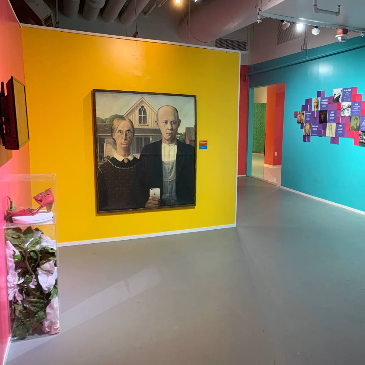 A brightly colored gallery space featuring a famous painting of a man and woman with a farmhouse, alongside a wall displaying colorful panels with text.