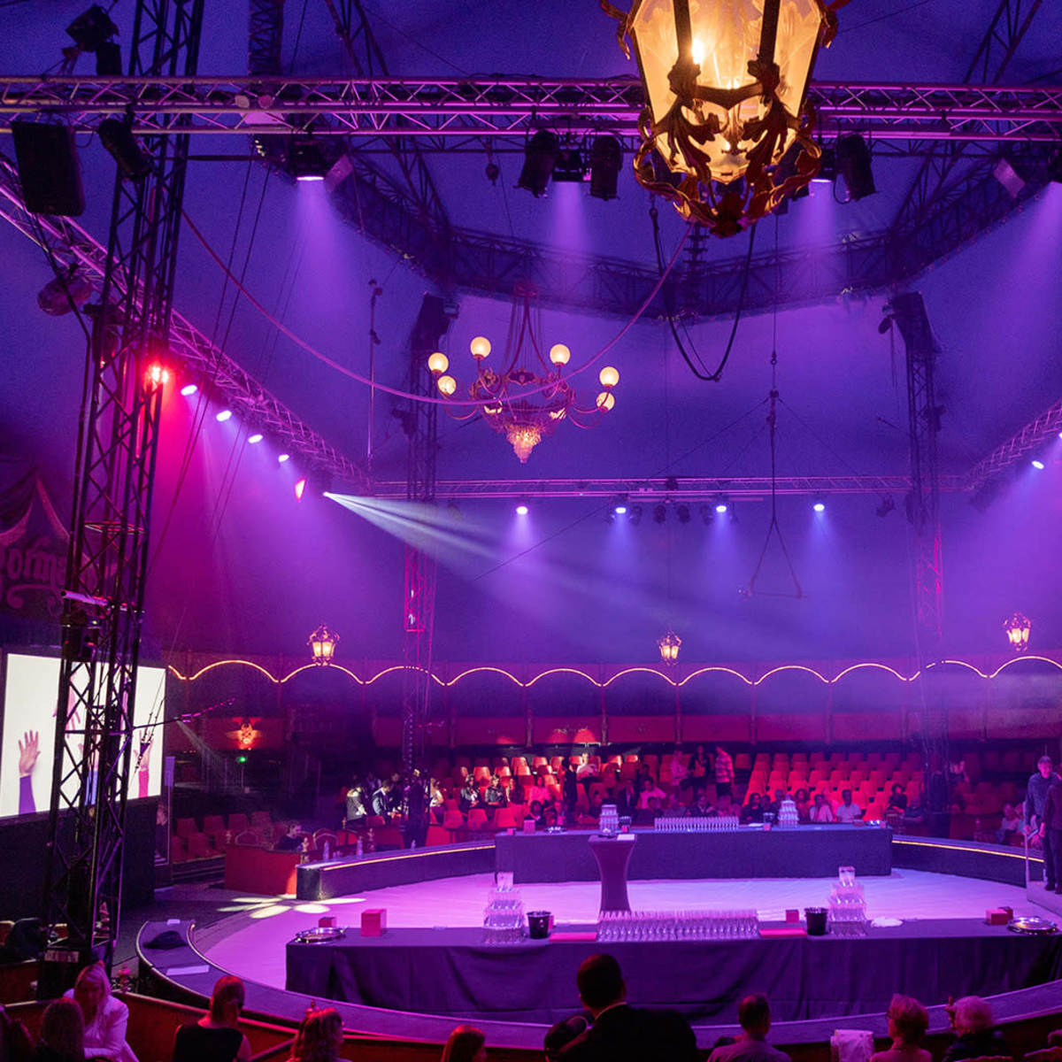 A circus tent interior features a central performance stage surrounded by seating and illuminated by colorful lights and chandeliers.
