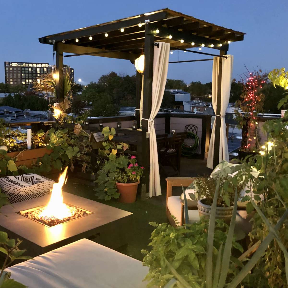 A rooftop terrace adorned with plants, comfortable seating, and a fire pit, illuminated by string lights at dusk.