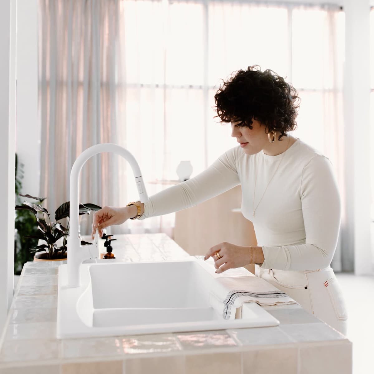 A woman with curly hair adjusts the faucet at a modern kitchen sink in a bright, airy room.