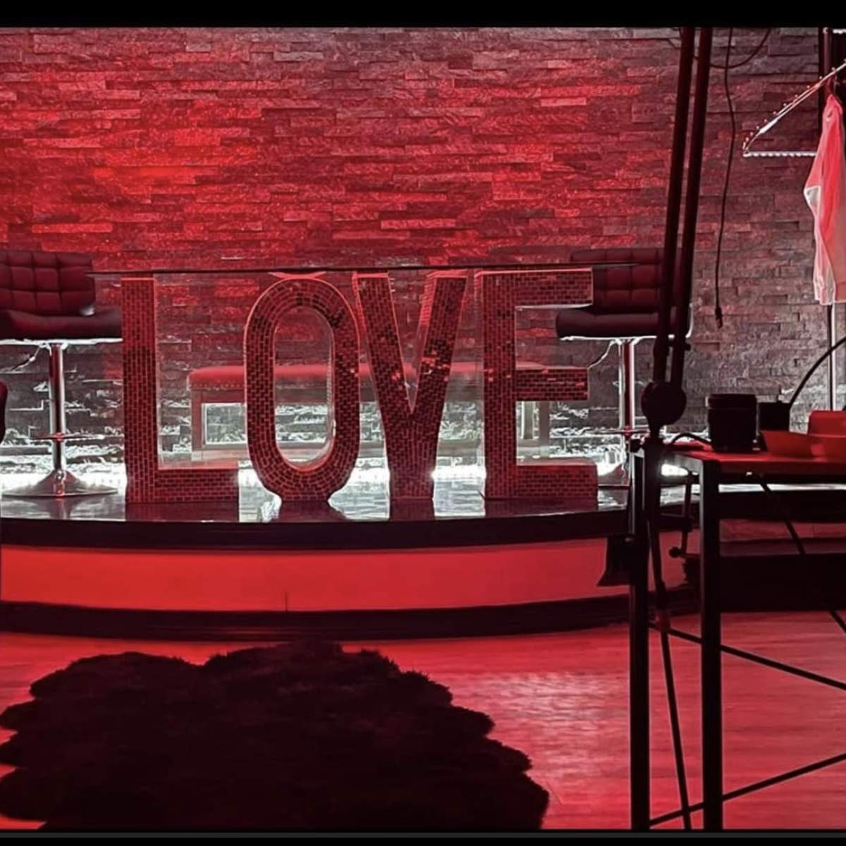 A red-lit interior with large, illuminated letters spelling "LOVE" on a stage-like setup, surrounded by modern chairs and a decorative rug.