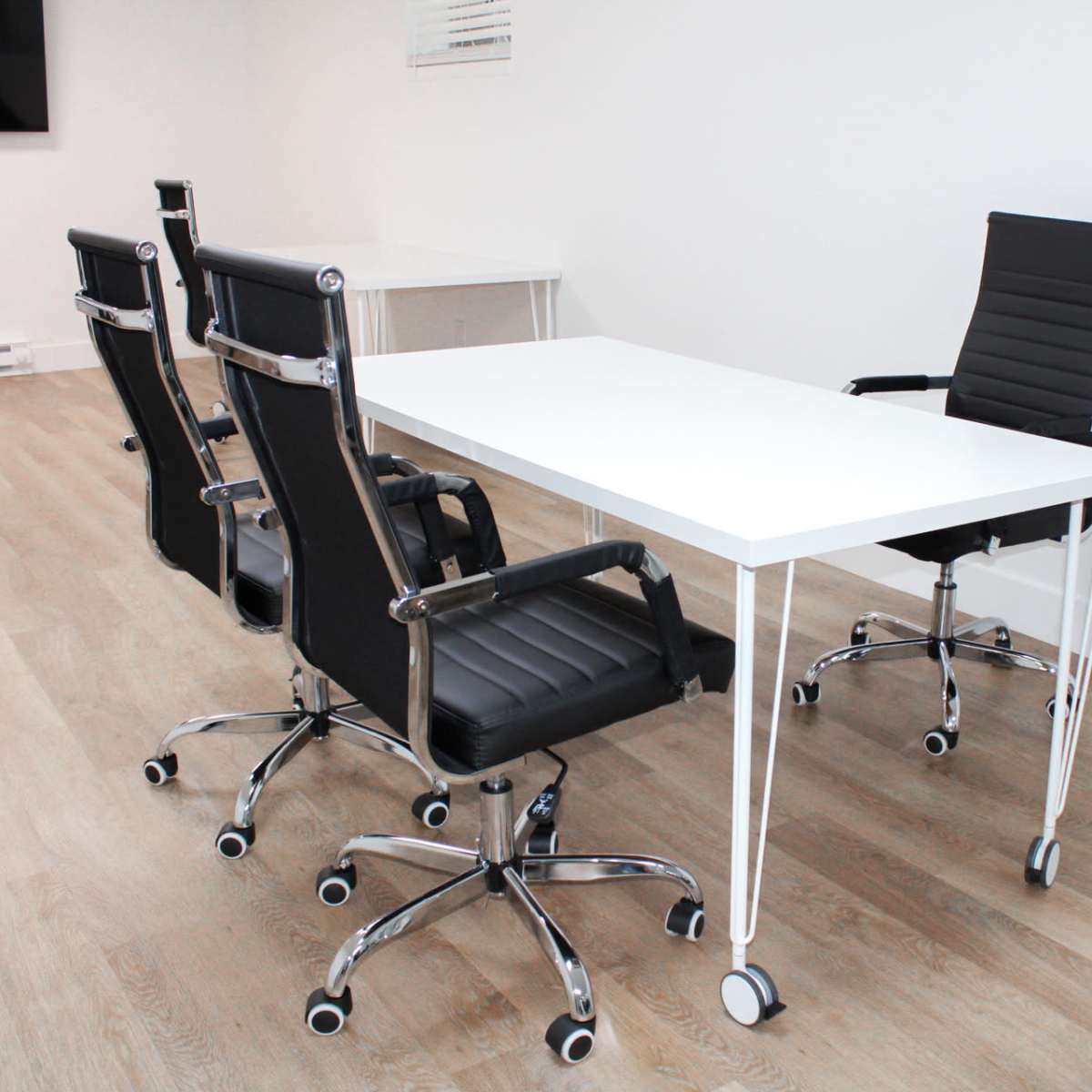 A modern conference room features a white table surrounded by four black office chairs on a light wood floor.