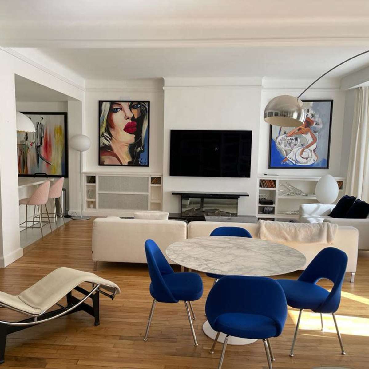 A modern living space features a marble dining table surrounded by blue chairs, a white sofa, and artwork on the walls, with a large TV and a fireplace in the background.