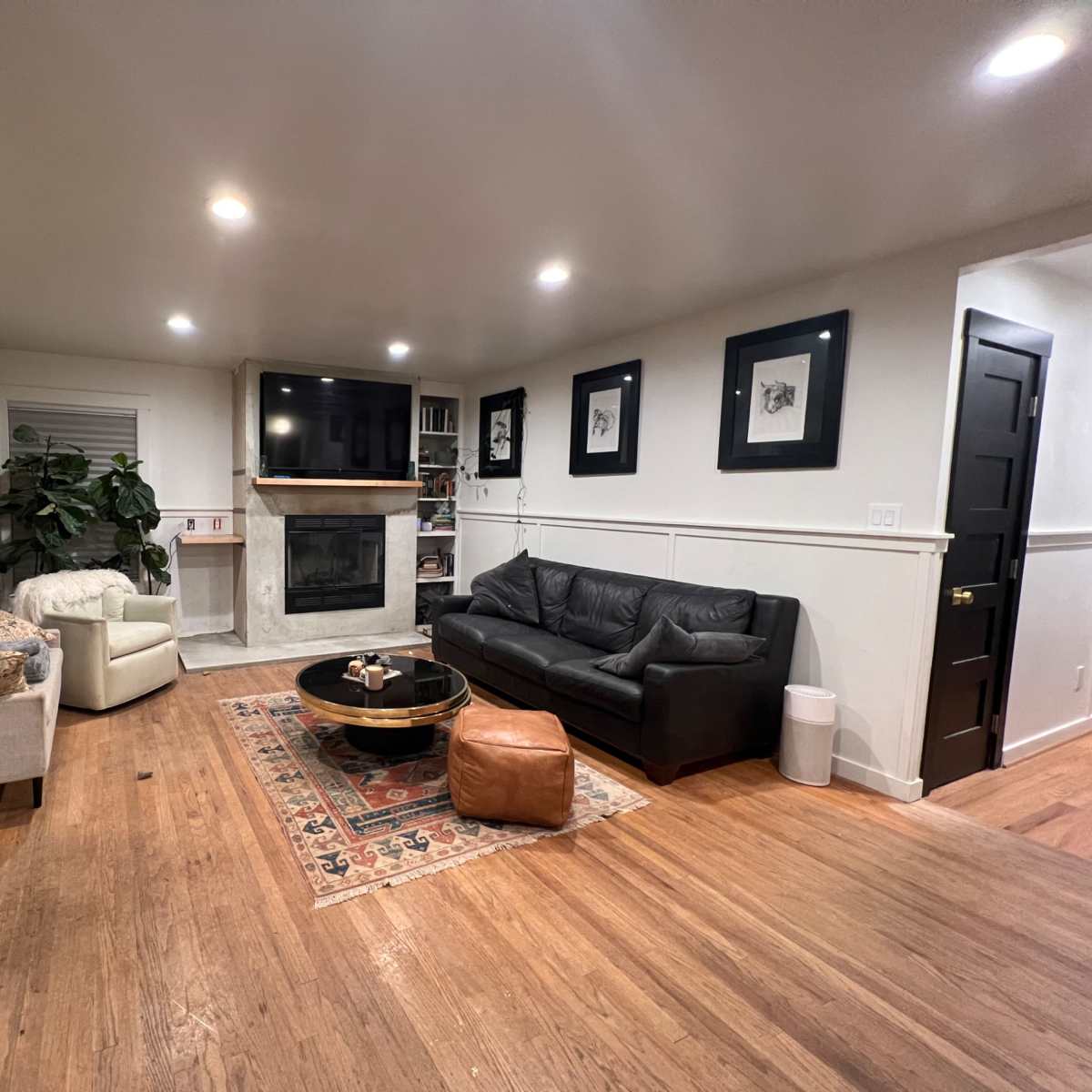 The living room features a leather sofa, a round coffee table, a flat-screen TV above a fireplace, and light wood flooring with a decorative rug.