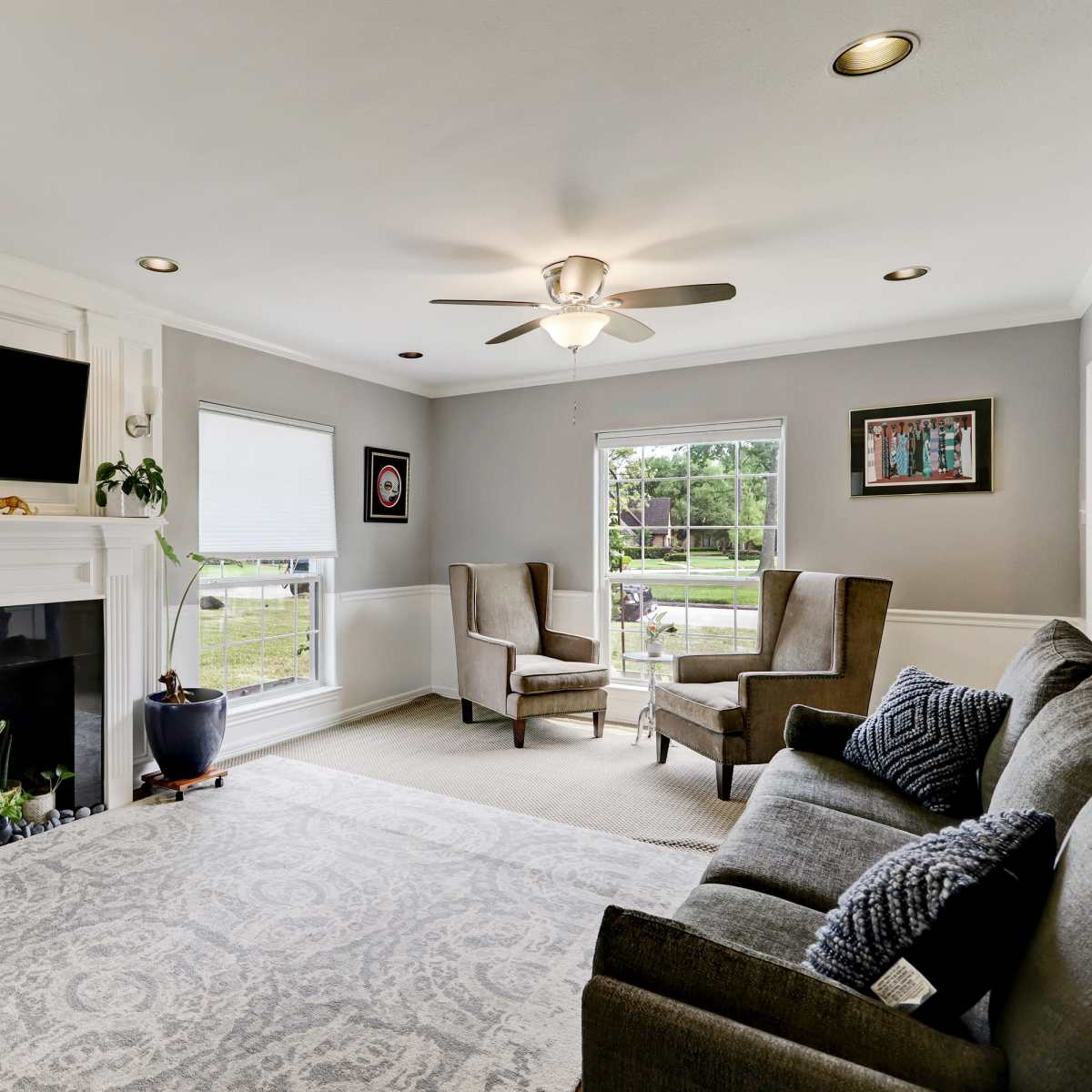 The living room features a pair of armchairs facing a couch, a fireplace with a television above it, and large windows allowing natural light to flood the space.