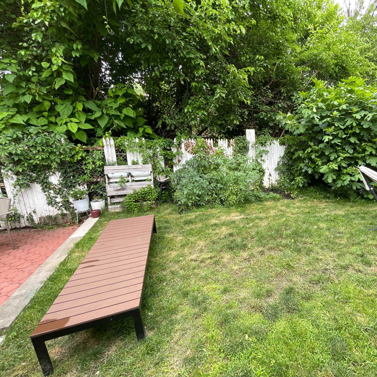 A small garden area with a grassy lawn, a wooden bench, a hammock, and lush green plants surrounded by a white wooden fence.