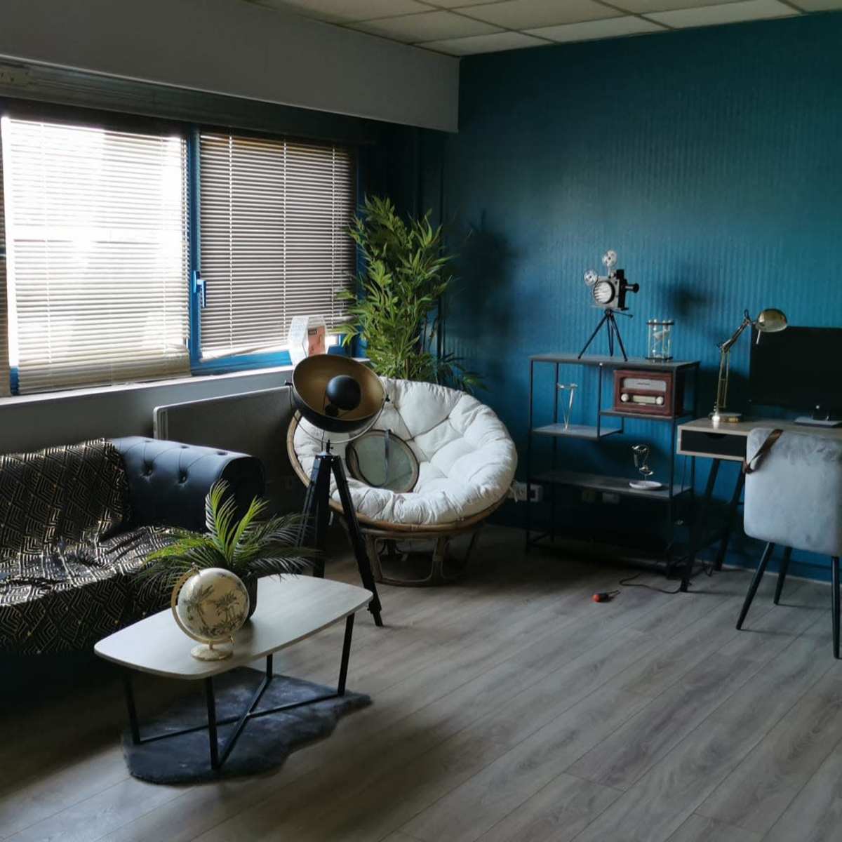 A modern, stylish room featuring a dark sofa, a round armchair, a small coffee table, a desk with a computer, and decorative plants against a teal wall.