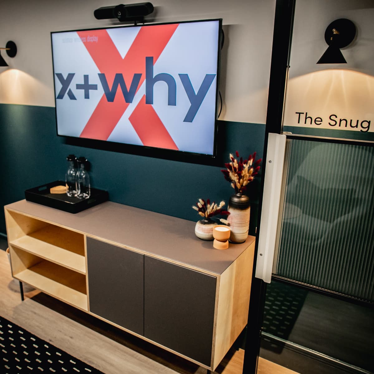 A modern room with a television displaying "x+why," a wooden cabinet holding decorative items, and stylish wall lighting.