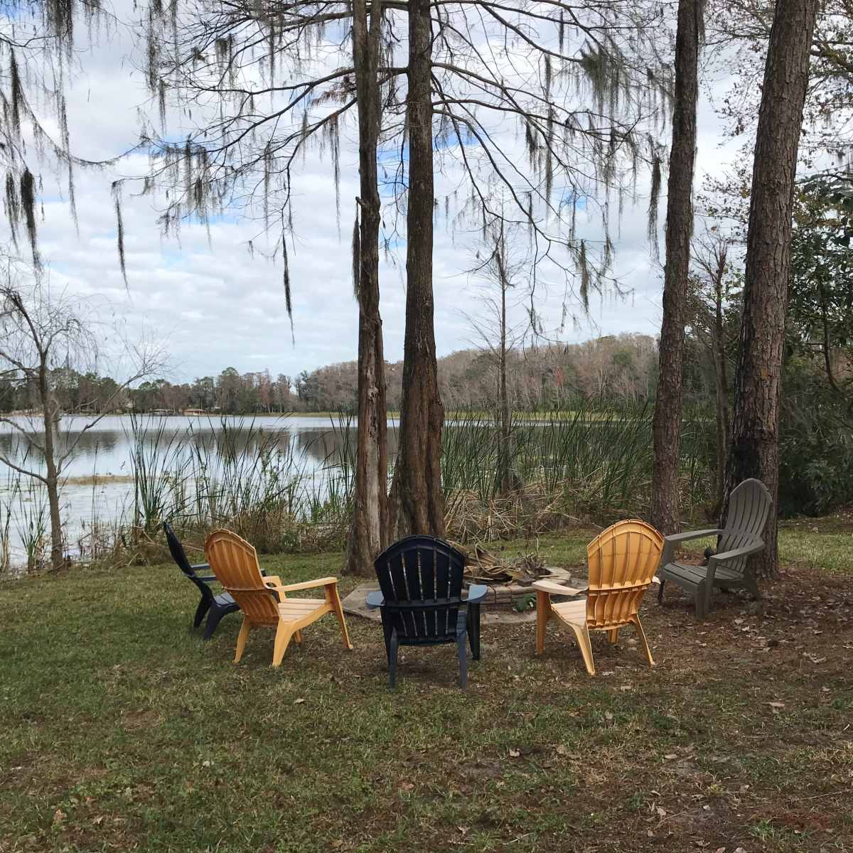 A circular arrangement of five outdoor chairs is situated near a calm lake surrounded by greenery and trees.