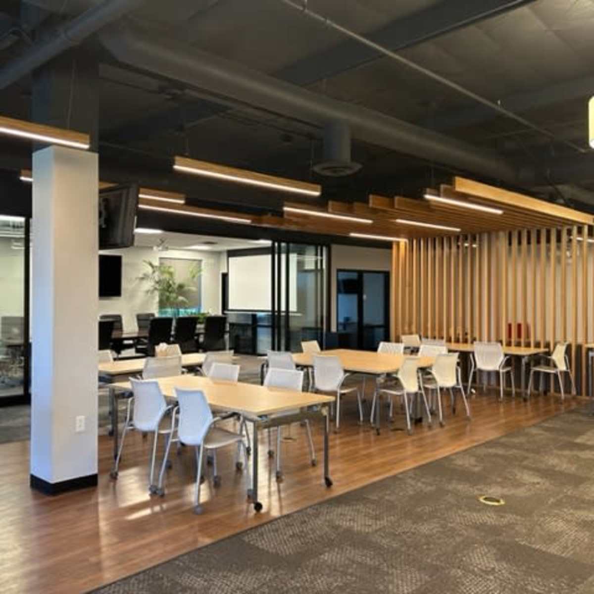 An office space with multiple tables and chairs, separated by wooden slat partitions, and glass-walled meeting rooms in the background.