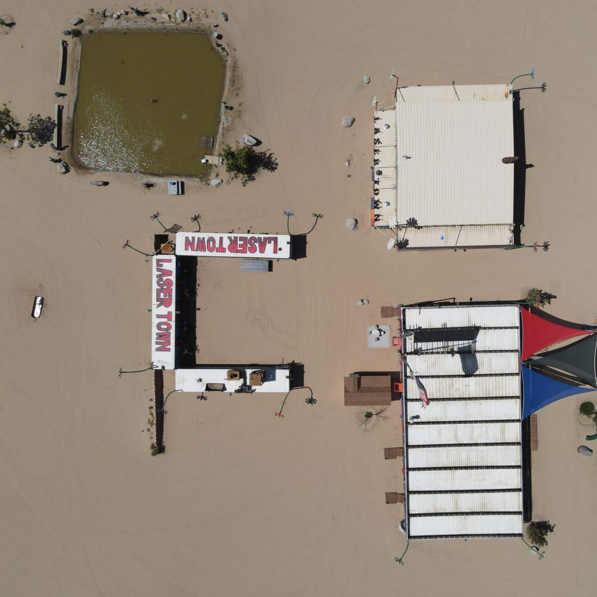 An aerial view of a desert landscape featuring several structures, including a building labeled "LASERTOWN," a pond, and a multi-roofed building.