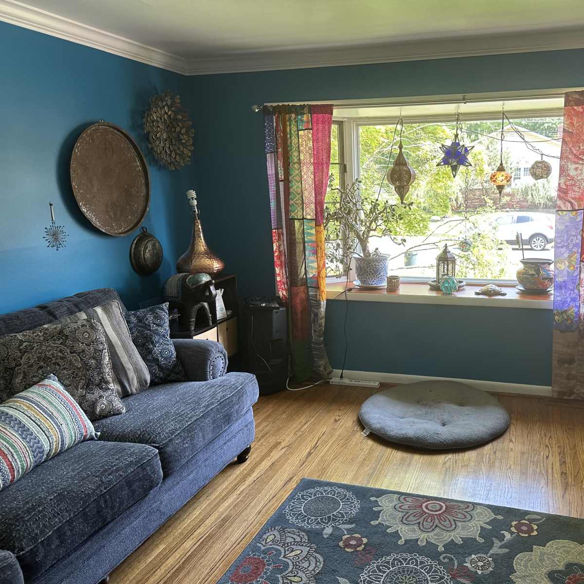 A living room with a blue wall, a sofa, colorful curtains, and a window displaying plants and a view of the outdoors.