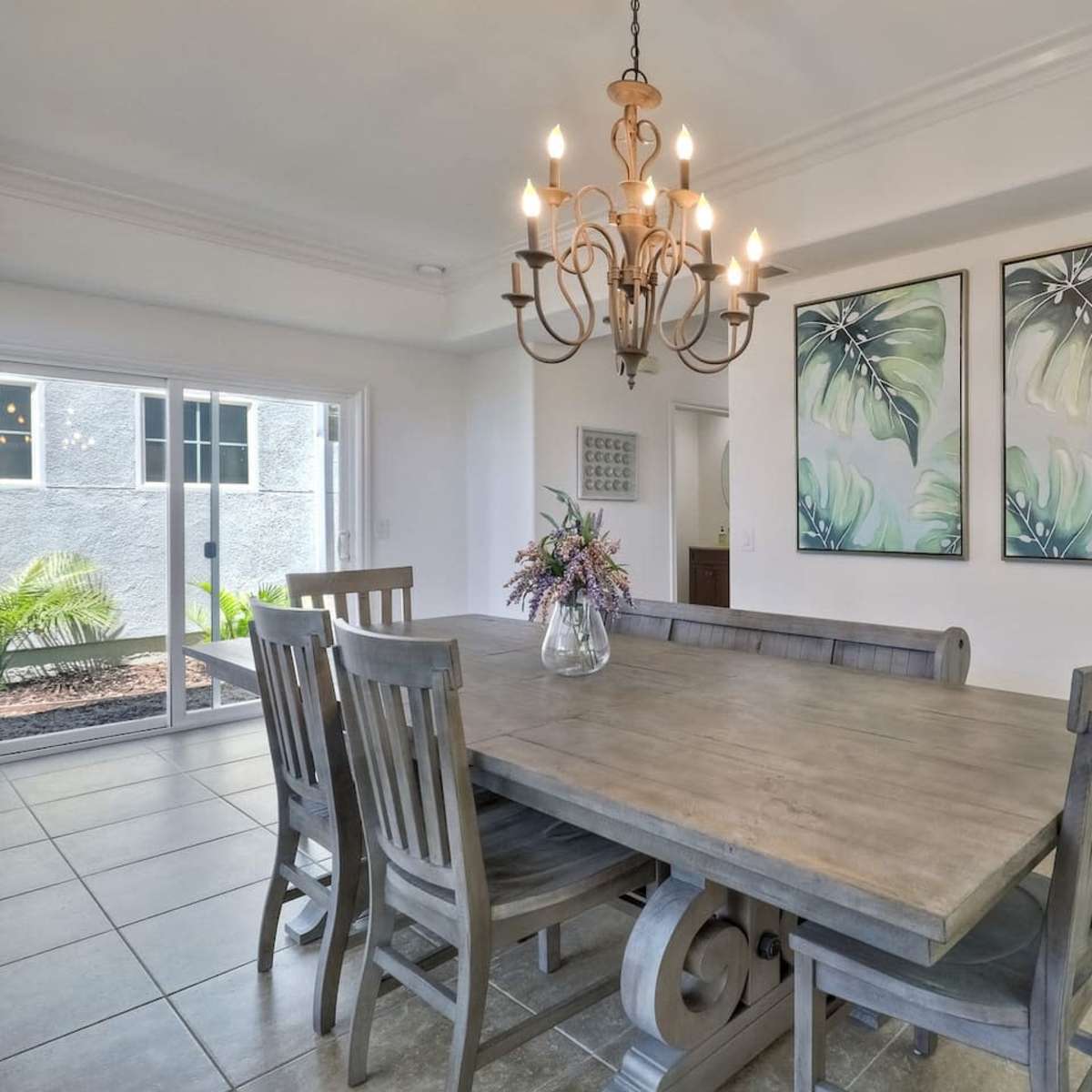 A dining area with a wooden table surrounded by six chairs, a chandelier above, and two framed botanical prints on the wall.