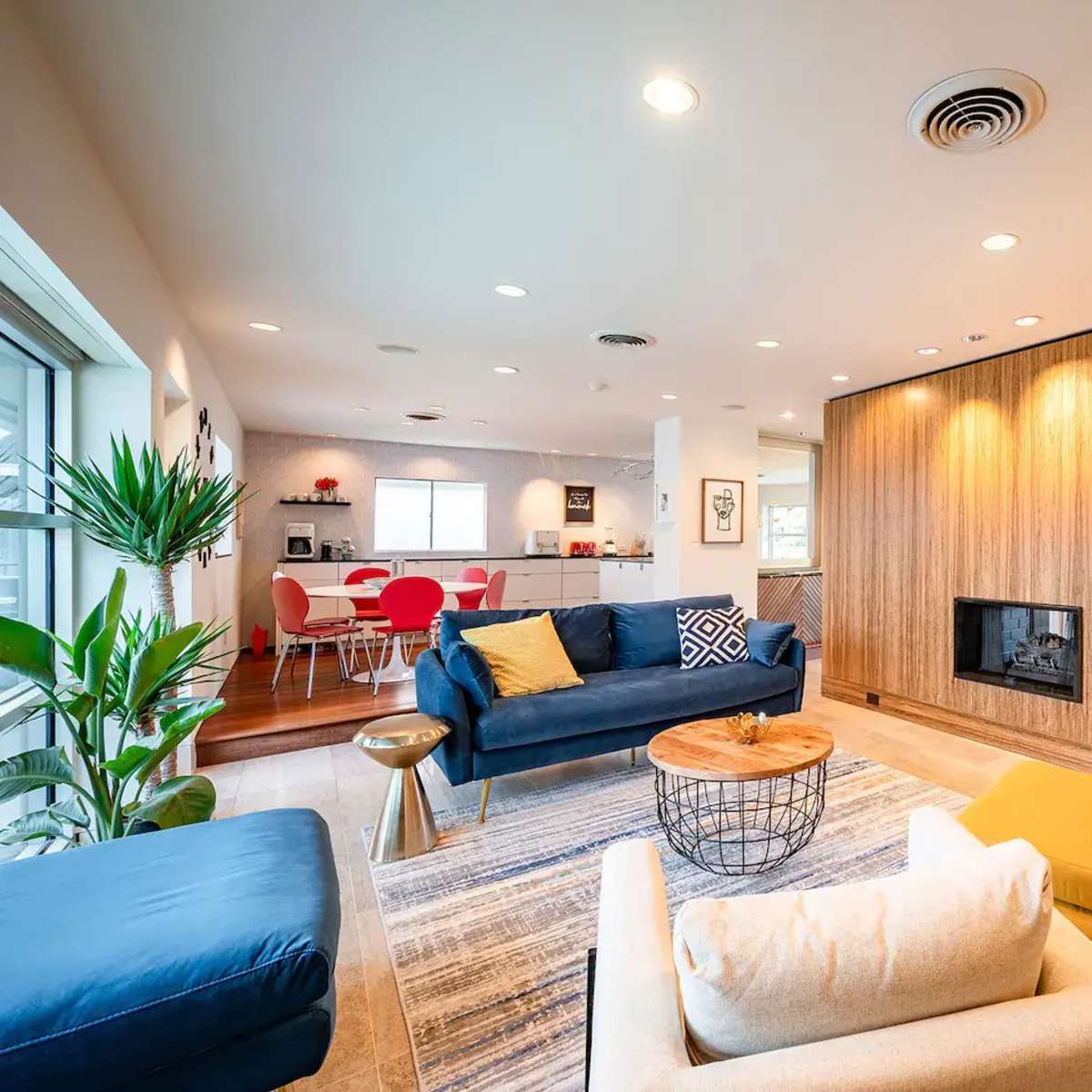 A modern living room features two sofas, a coffee table, and a wall-mounted fireplace, with a dining area visible in the background.