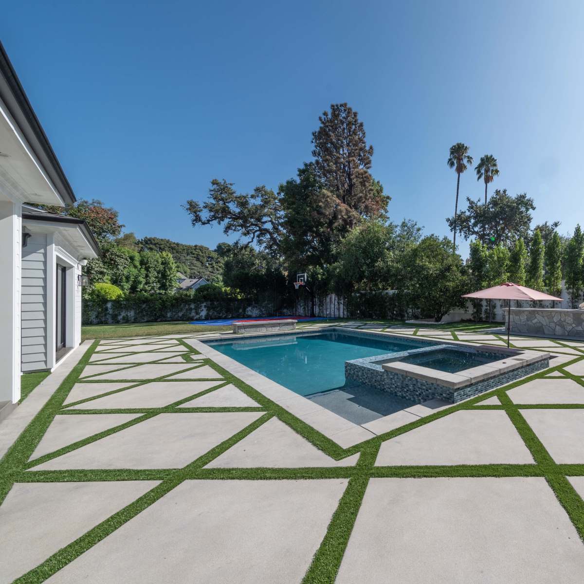 A geometric concrete patio with a swimming pool and a landscaped yard featuring trees and an umbrella.