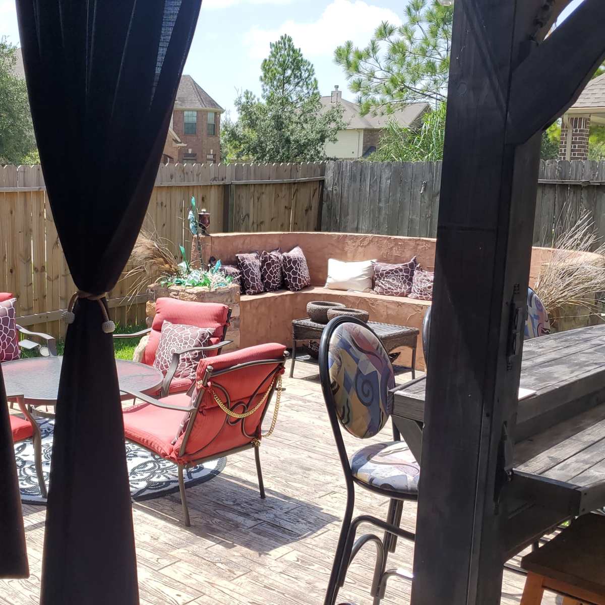 A backyard patio area with seating arrangements, a table, and decorative plants enclosed by a wooden fence.