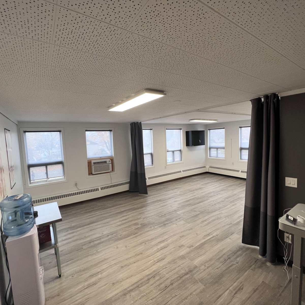 An empty room with wooden flooring, multiple windows, a wall-mounted television, and medical posters hanging on the walls.