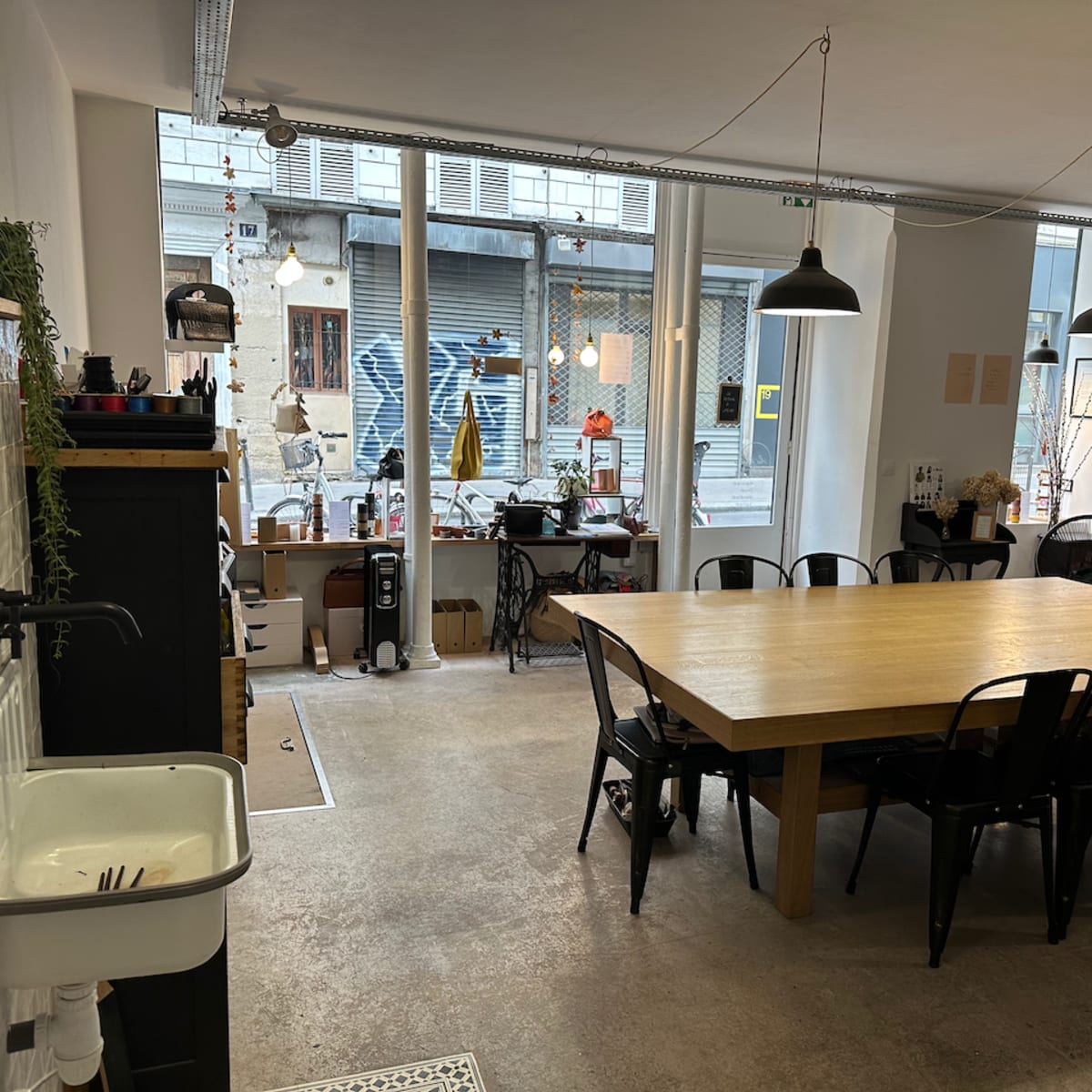 A spacious, modern kitchen area features a large wooden table surrounded by black chairs, with large windows showing an urban street outside.
