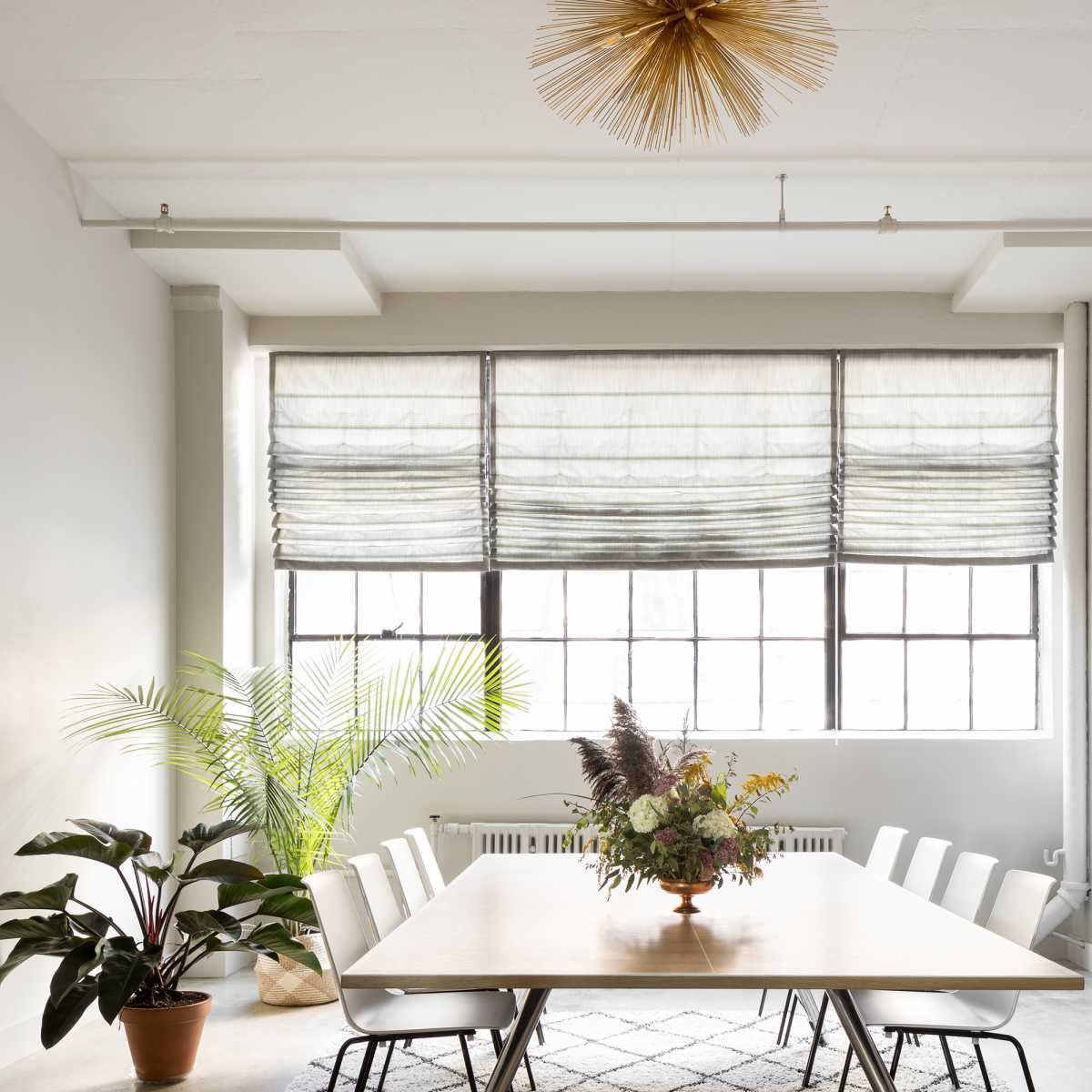 A modern dining room features a large table surrounded by white chairs, a decorative plant in the corner, and a television on a low cabinet beneath large windows with textured shades.