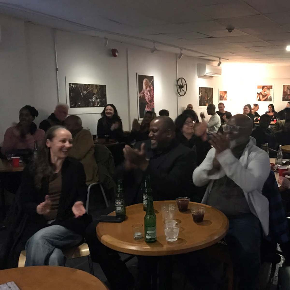 A diverse crowd of people sits at tables in a warmly lit venue, clapping and smiling during a performance.