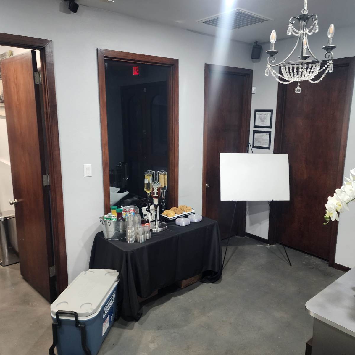 A small event space with two wooden doors, a table covered with a black cloth displaying drinks and snacks, and a blank easel on display.