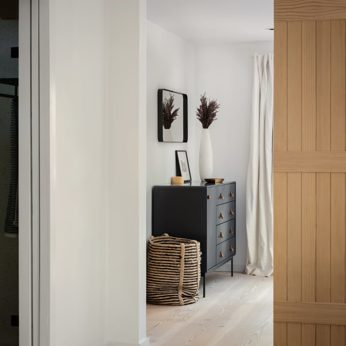 A hallway with a wooden sliding door leads to a room featuring a black dresser, a decorative vase, and a woven basket.