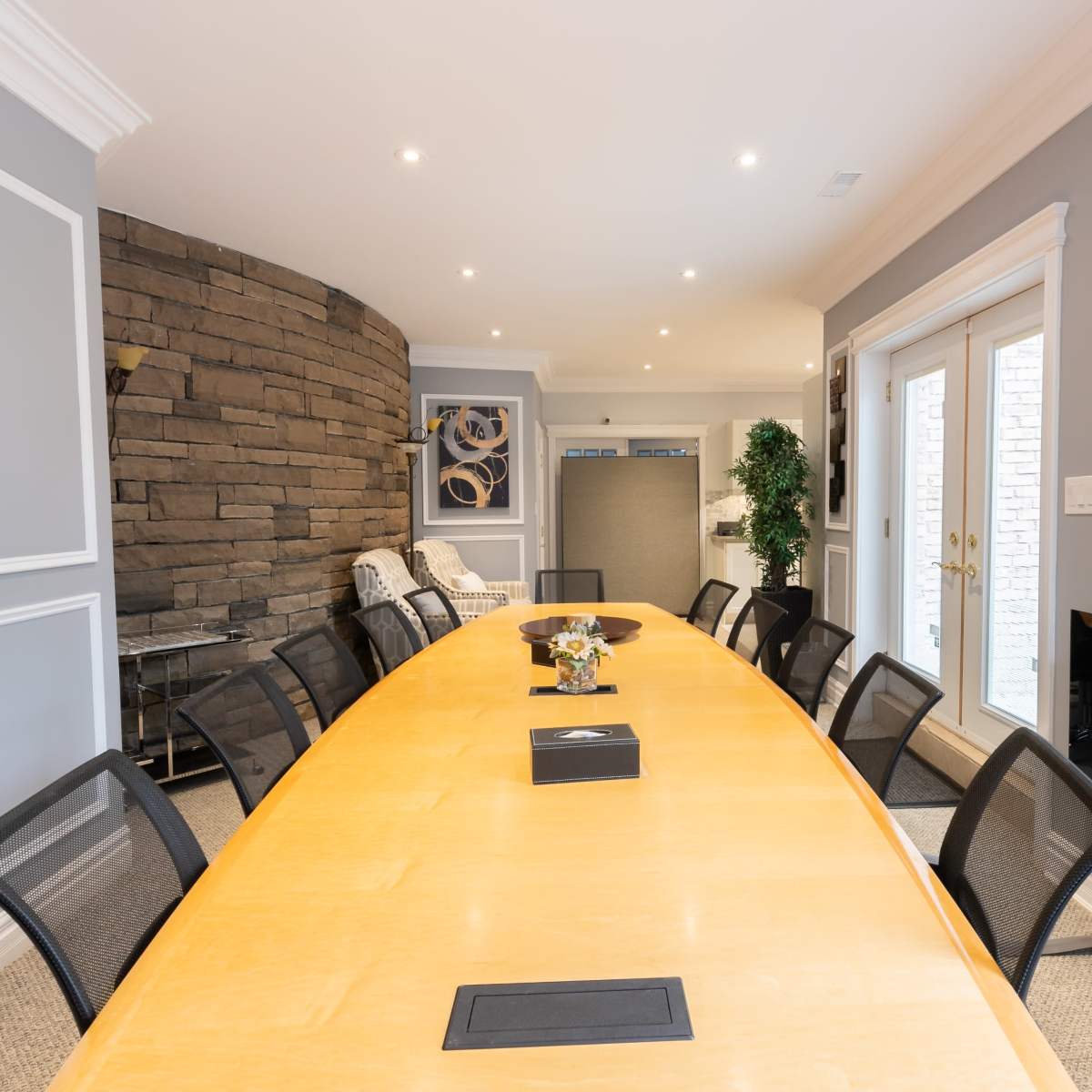 A long wooden conference table is surrounded by black chairs in a well-lit meeting room with artwork and a stone wall in the background.