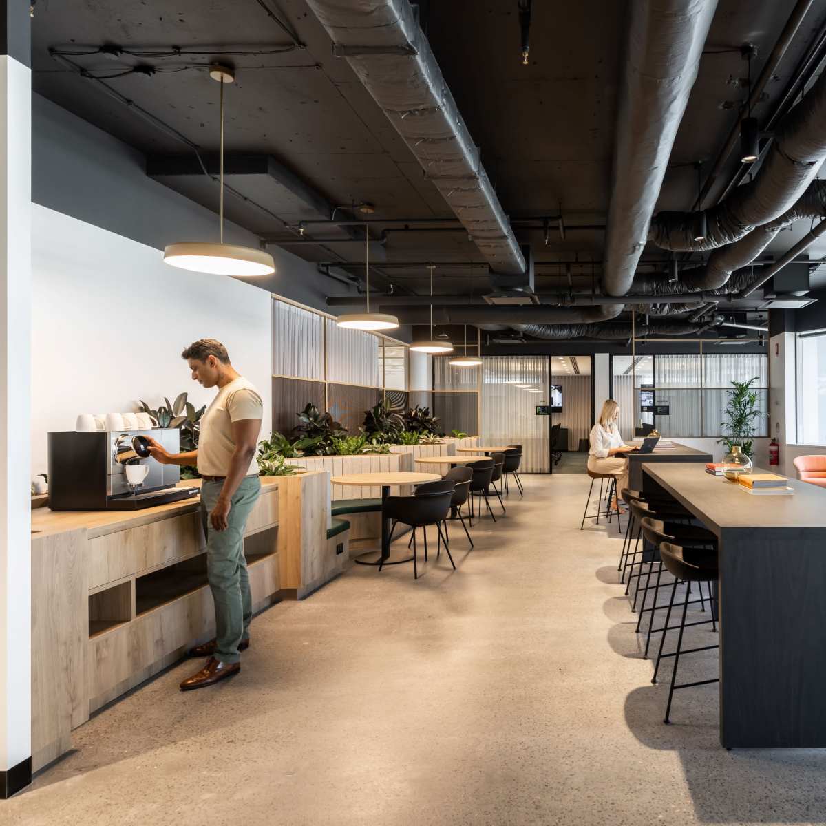 An office breakroom features a coffee station, seating areas, and large windows surrounded by plants.