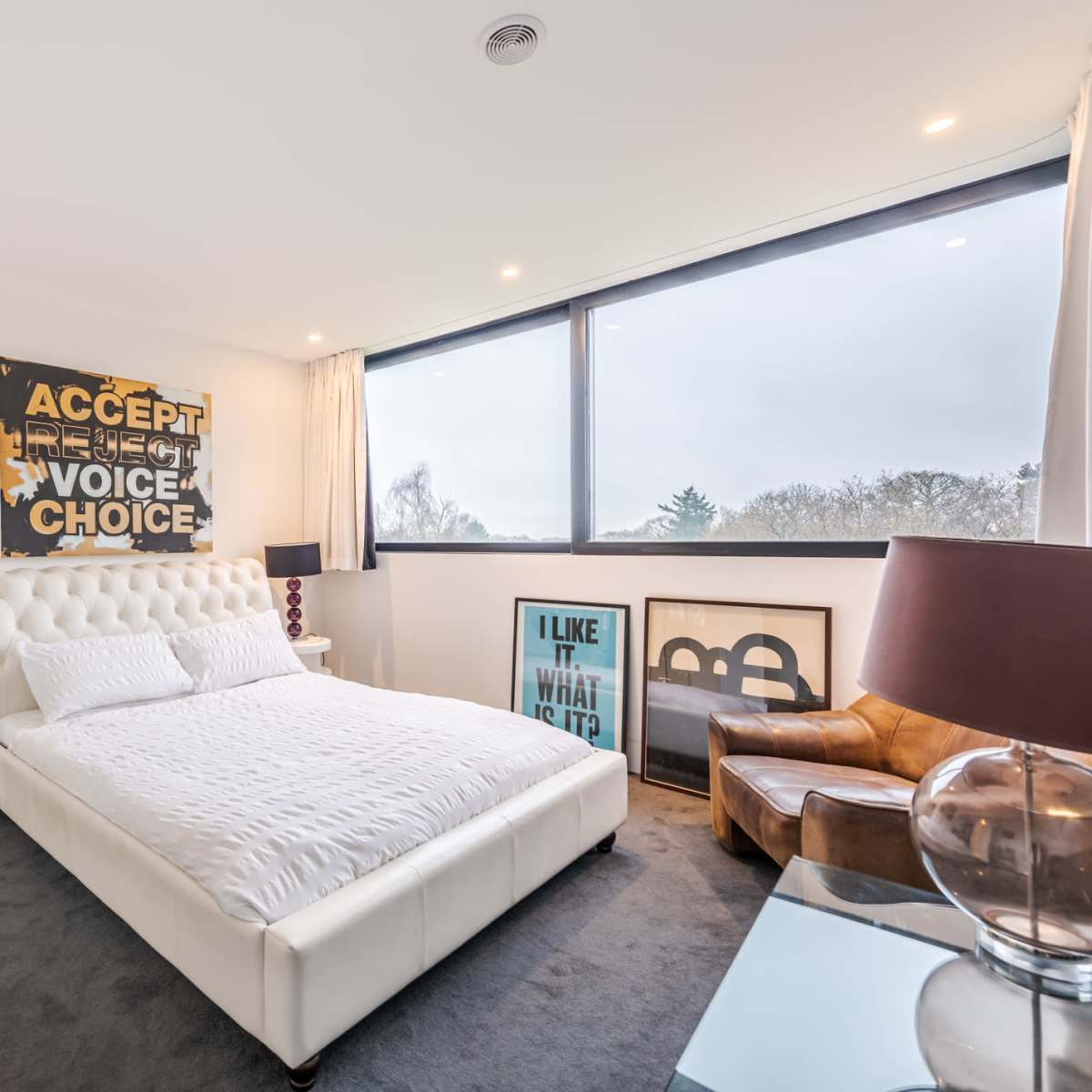 A modern bedroom features a large white bed, a brown leather chair, and artwork on the walls, illuminated by natural light from a large window.