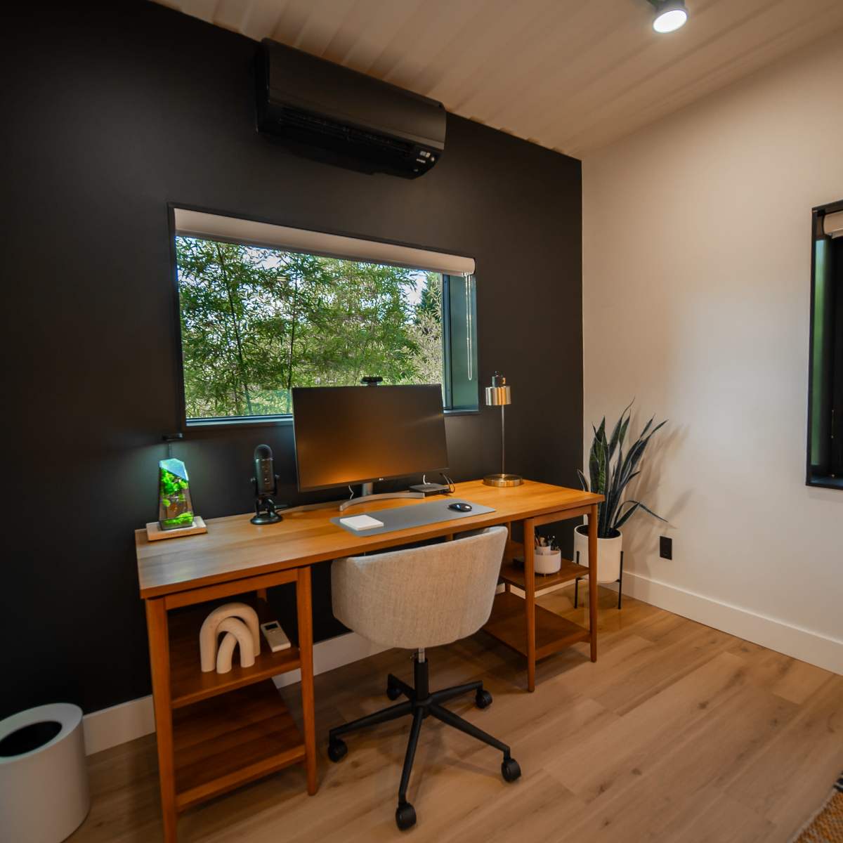 A modern home office with a wooden desk, a computer monitor, and a chair, situated next to a window overlooking greenery.