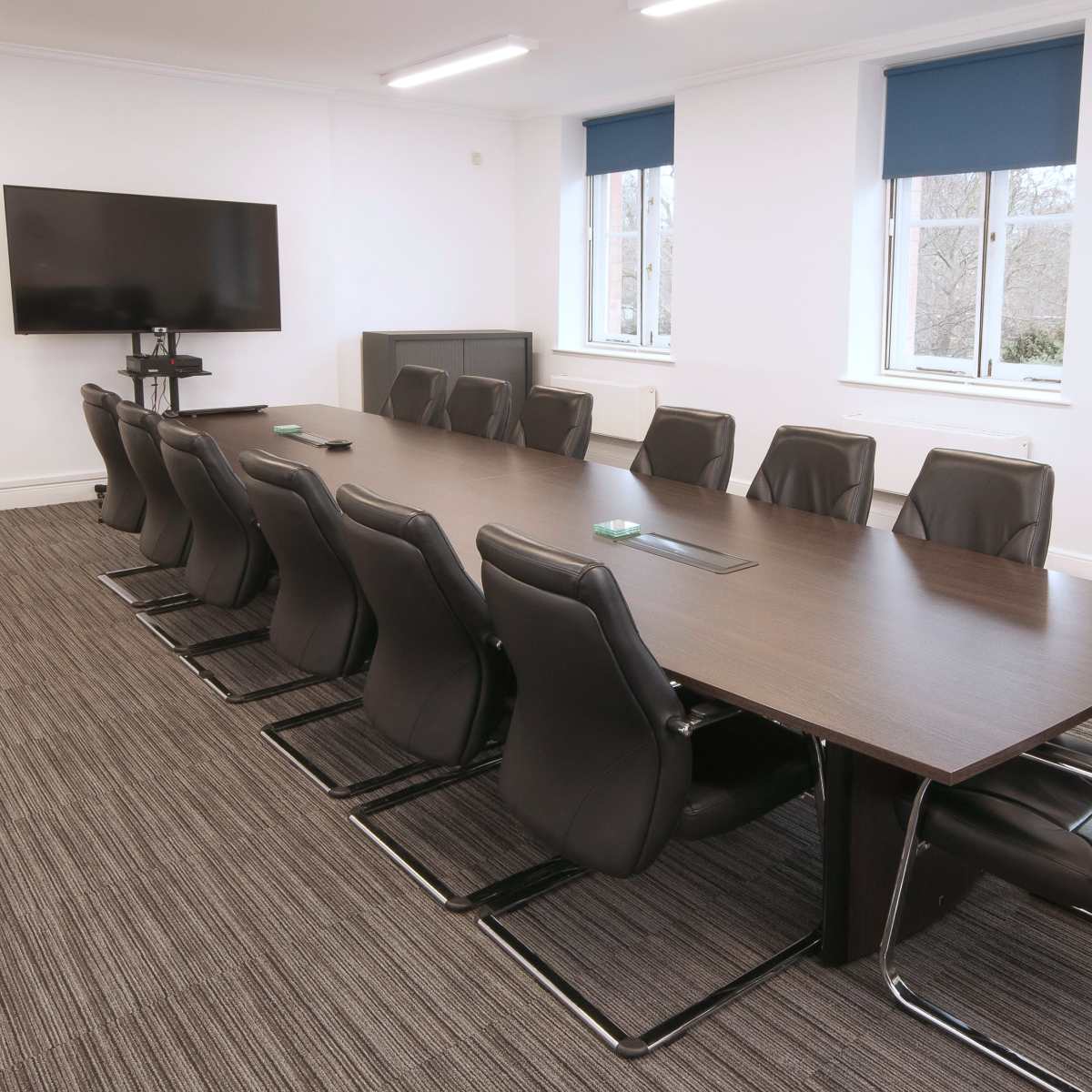 A long conference table with black leather chairs is set up in a bright, modern meeting room featuring a large wall-mounted screen and windows with blue shades.