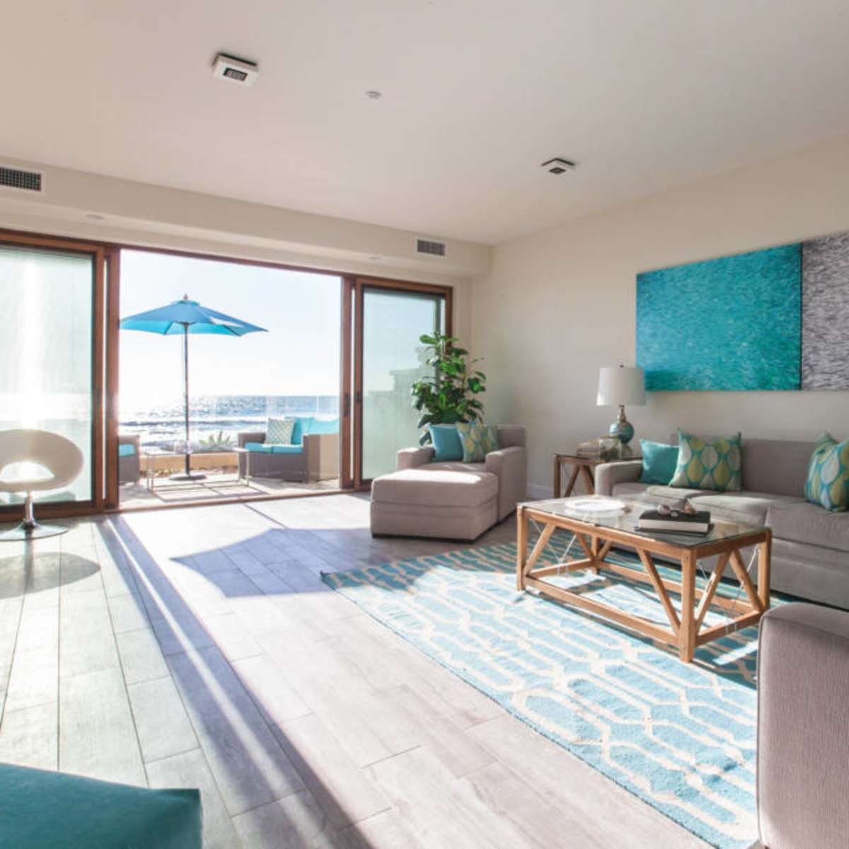 A modern living room with large windows overlooking the ocean, featuring two sofas, a coffee table, and an umbrella-covered outdoor seating area.