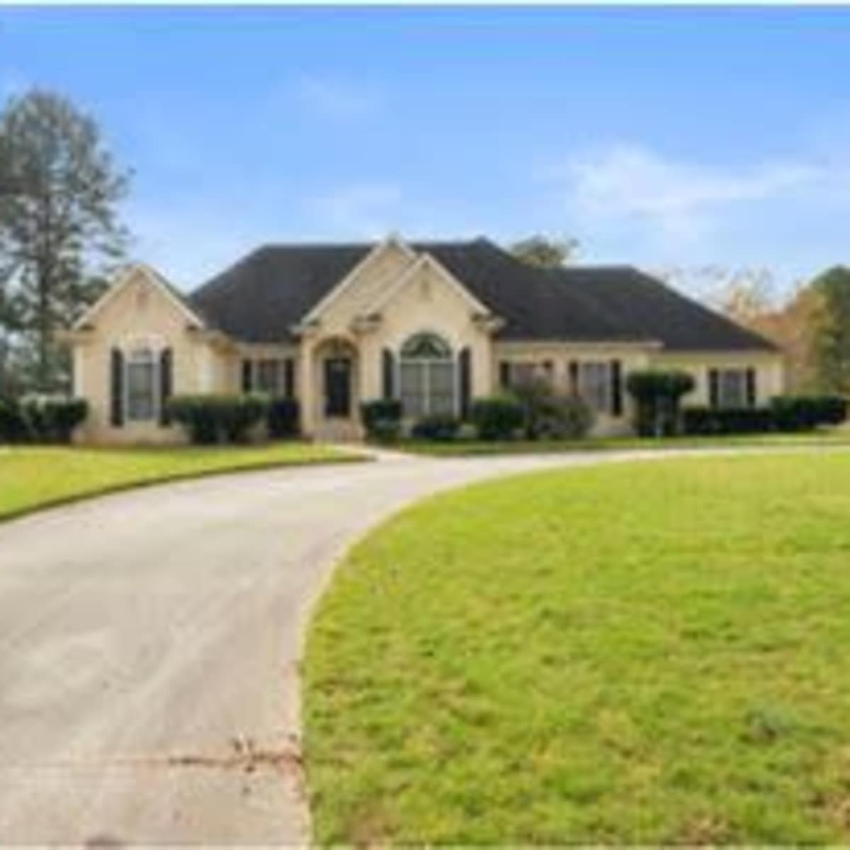 A single-story house with a front lawn and a curved driveway is surrounded by trees and a clear sky.