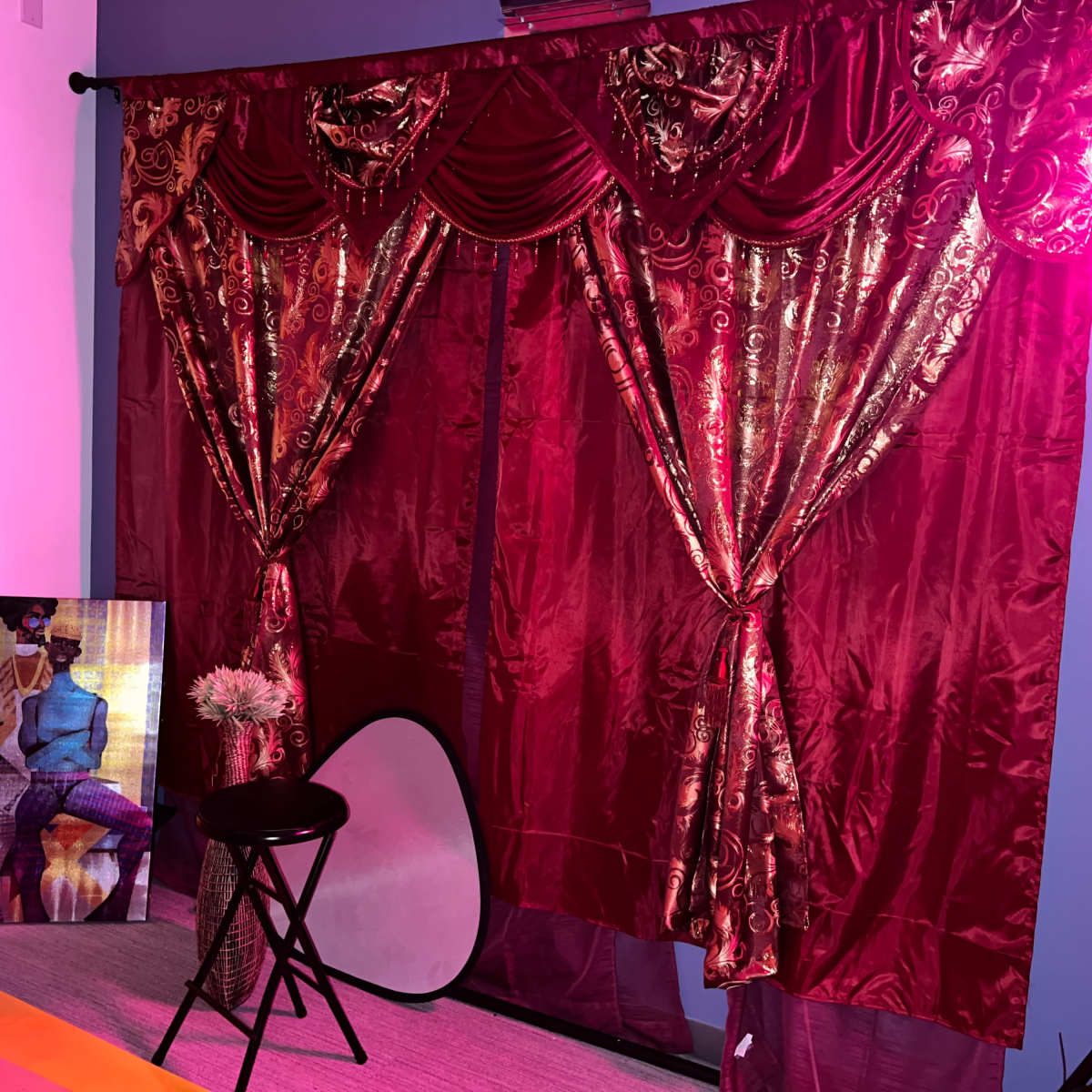 The scene features a backdrop of red curtains with ornate patterns, a framed artwork on the floor, a stool, and a reflector disk in a brightly lit room.