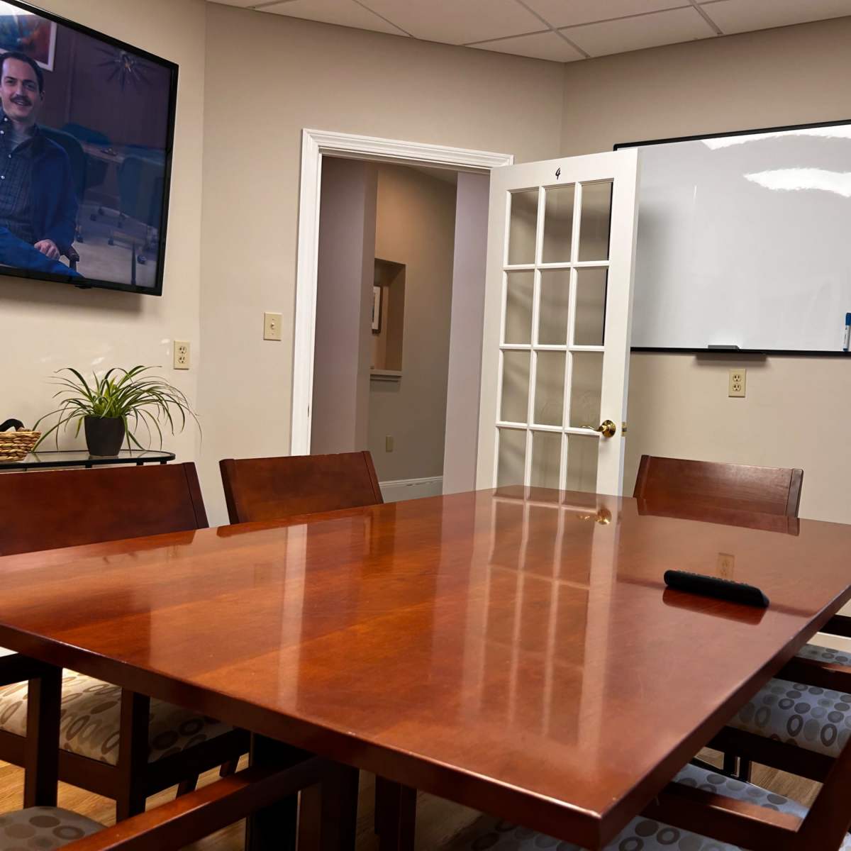 A wooden conference table with chairs is positioned in a meeting room, featuring a television on the wall and a whiteboard nearby.