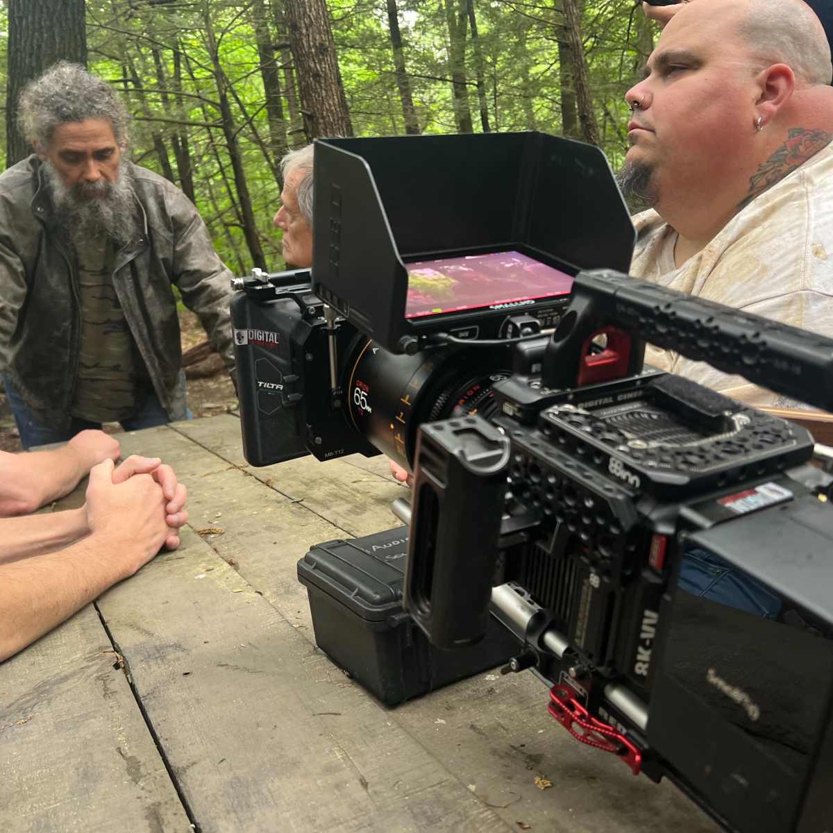 A film crew is gathered at a wooden table in a forest, with cameras focused on three men engaged in conversation.