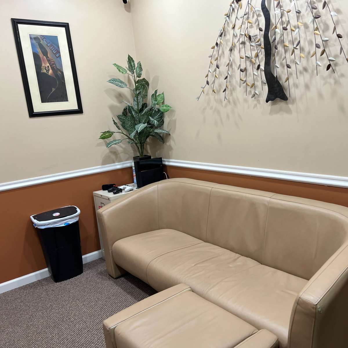 A small room with a tan couch, a trash can, a potted plant, and a wall decoration of a tree with leaves.