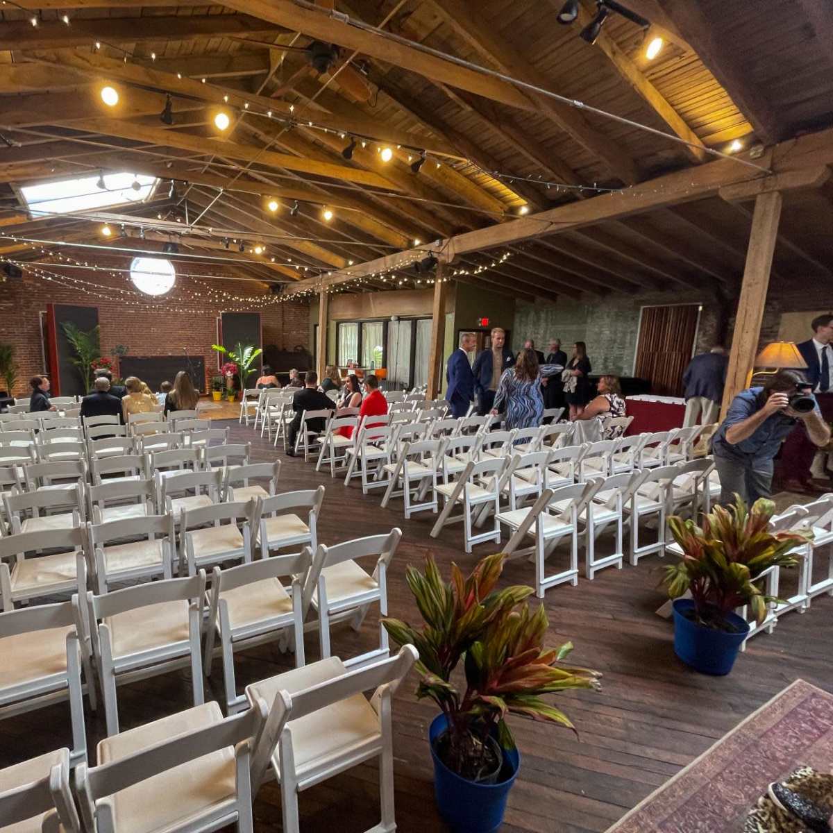 A spacious event venue with rows of white chairs set up for an audience, featuring wooden beams, string lights, and potted plants.