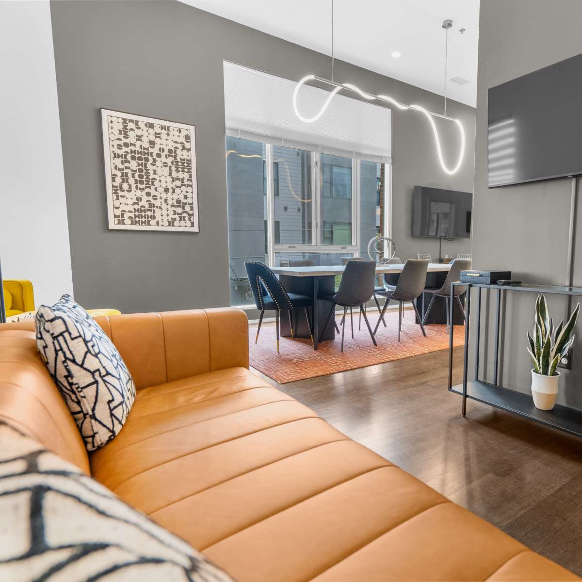 A modern living space features a brown leather sofa, a television mounted on the wall, and a dining area with a table and chairs visible in the background.