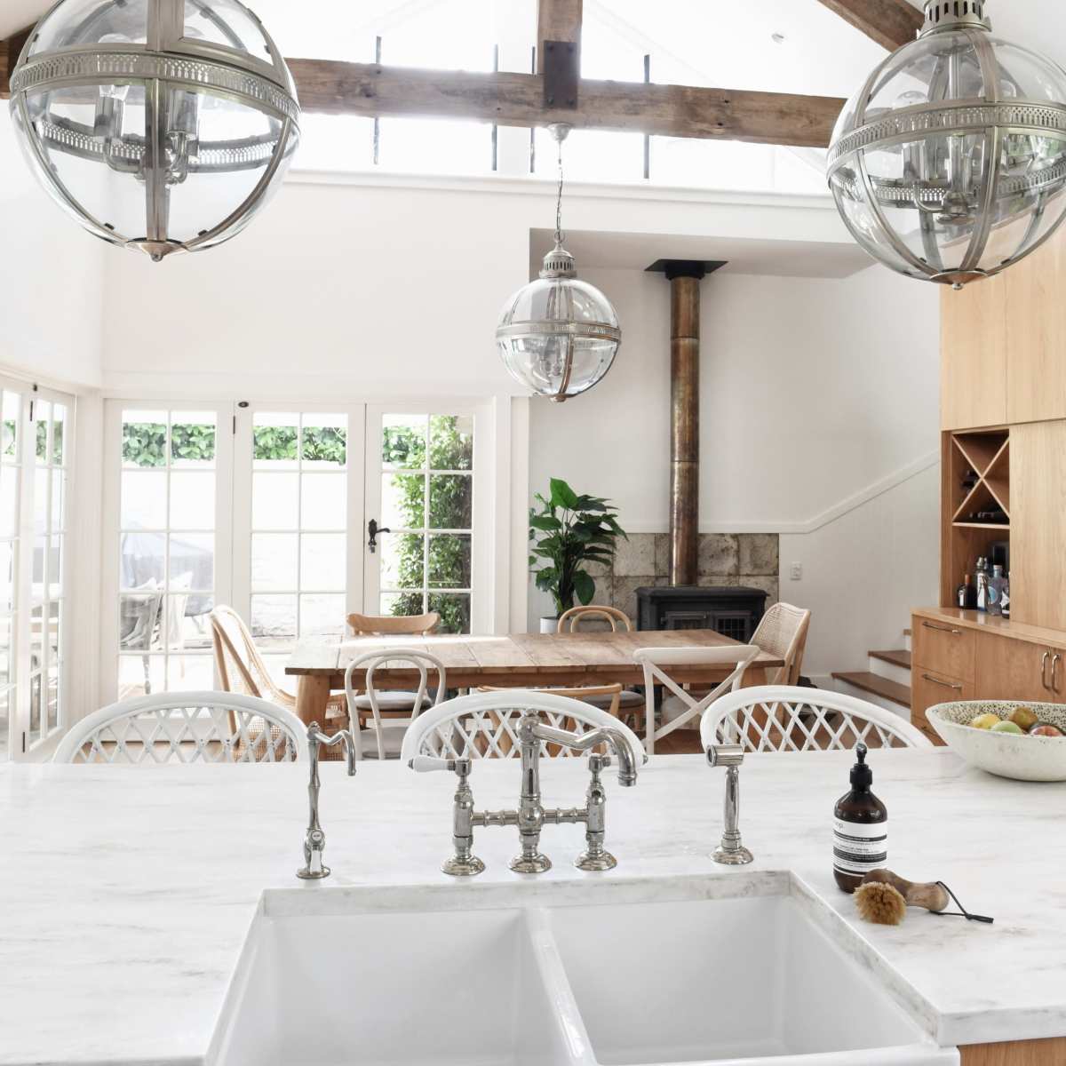 A bright kitchen with a large farmhouse sink, a wooden dining table in the background, and three pendant lights hanging from a vaulted ceiling.